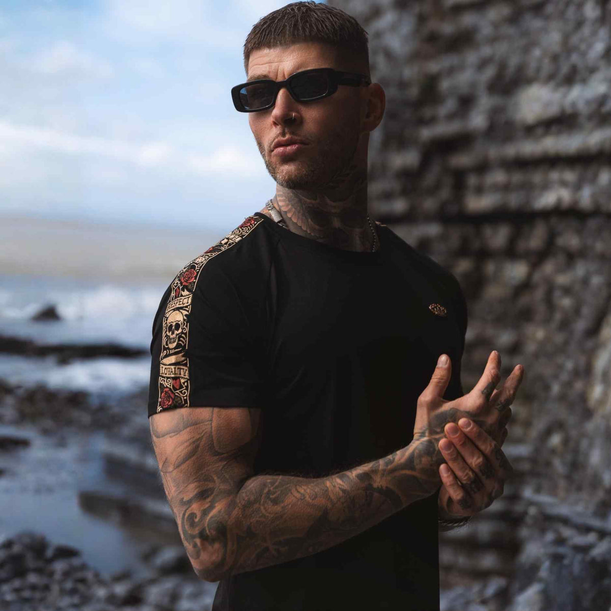 Model mit Sonnenbrille im schwarzen LFR T-Shirt mit farbigem Ärmelband an der Küste