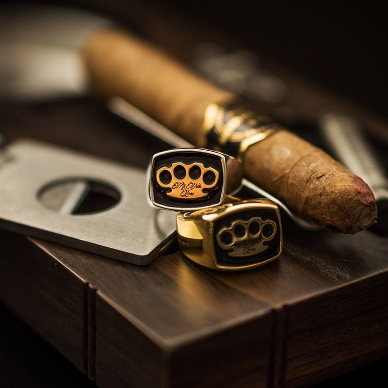 Anillos de sello MVL plateado y dorado junto a un puro y un cortapuros sobre madera